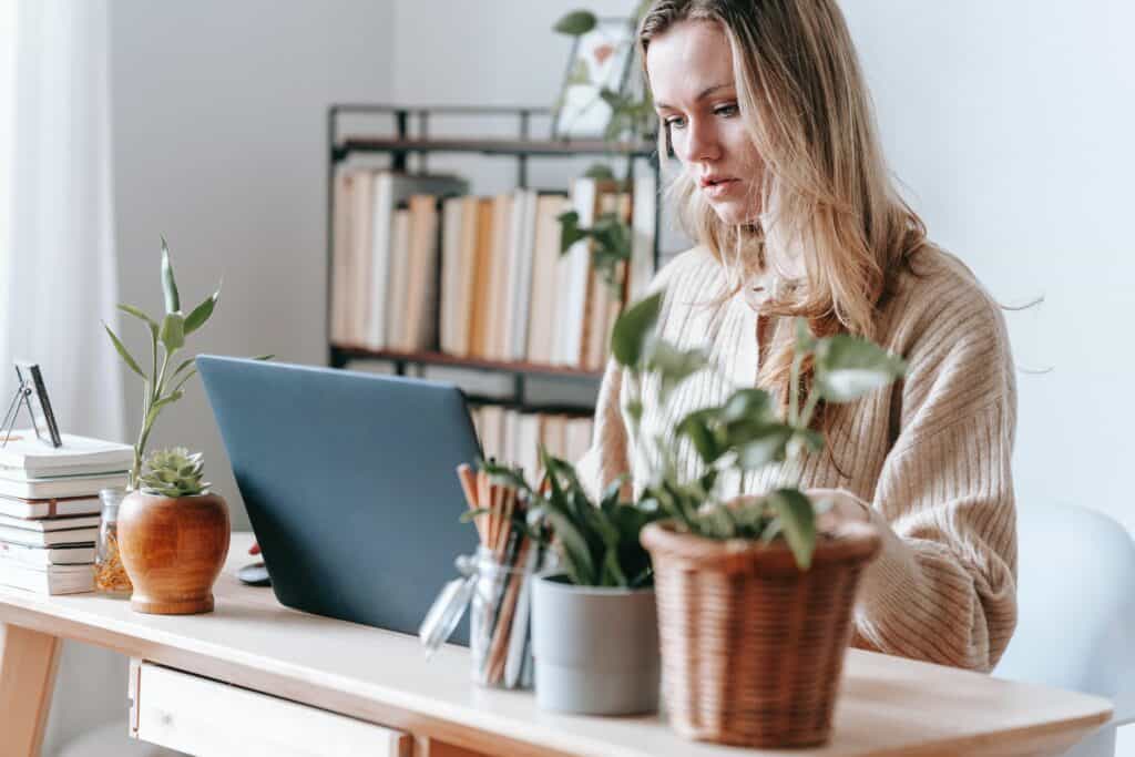 A person focuses on a laptop at a wooden desk adorned with potted plants and books, set in a bright room with a bookshelf in the background.