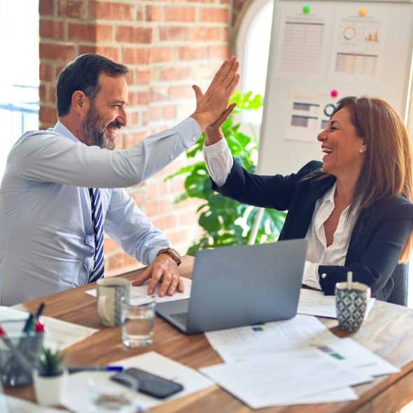 Two people in business attire high-five in an office, surrounded by documents, a laptop, and a brick wall. A whiteboard with charts and plants are in the background.